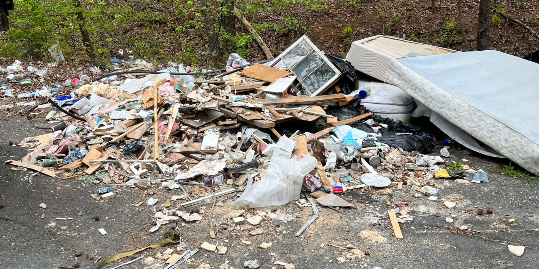 A pile of discarded items and debris, including a mattress and construction materials, is scattered across an outdoor area by a road. The surrounding area has sparse vegetation and trees.