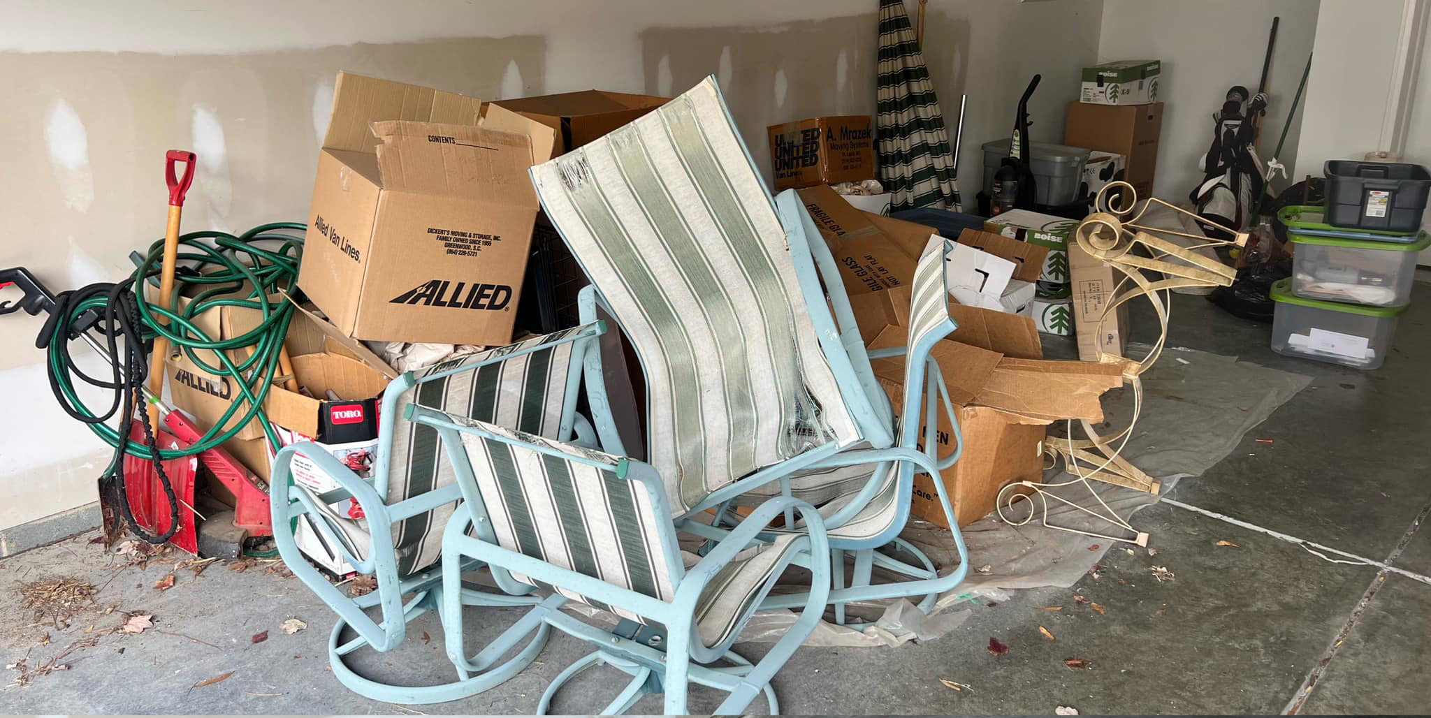 A cluttered garage corner with stacked cardboard boxes, two striped patio chairs, a beige metal plant stand, garden hoses, a red shovel, and various containers. The floor has scattered leaves and small debris.