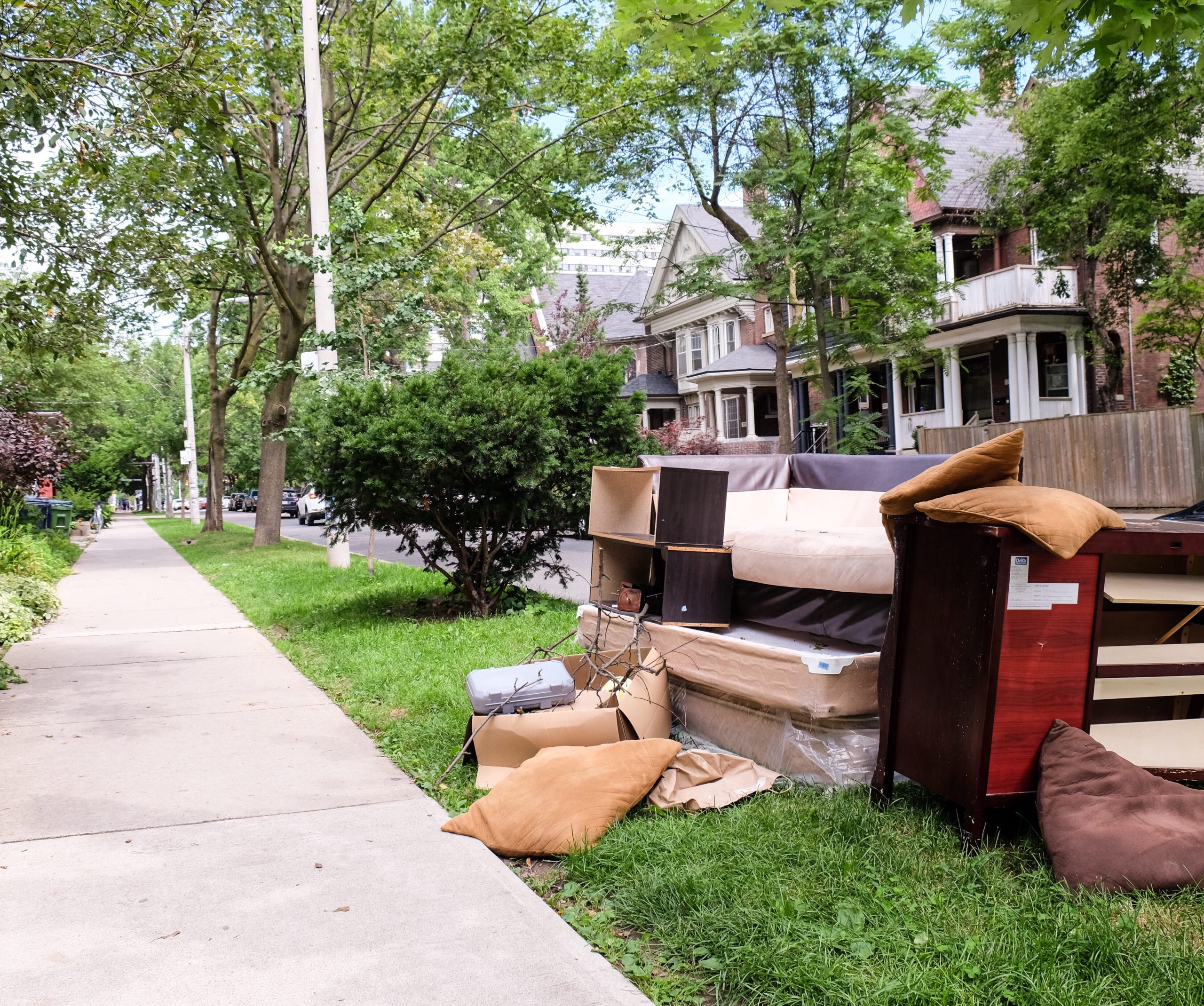 A pile of furniture, including a couch, dresser, and cushions, is placed on the grassy curbside in a residential neighborhood with tree-lined streets and vintage-style houses in the background.